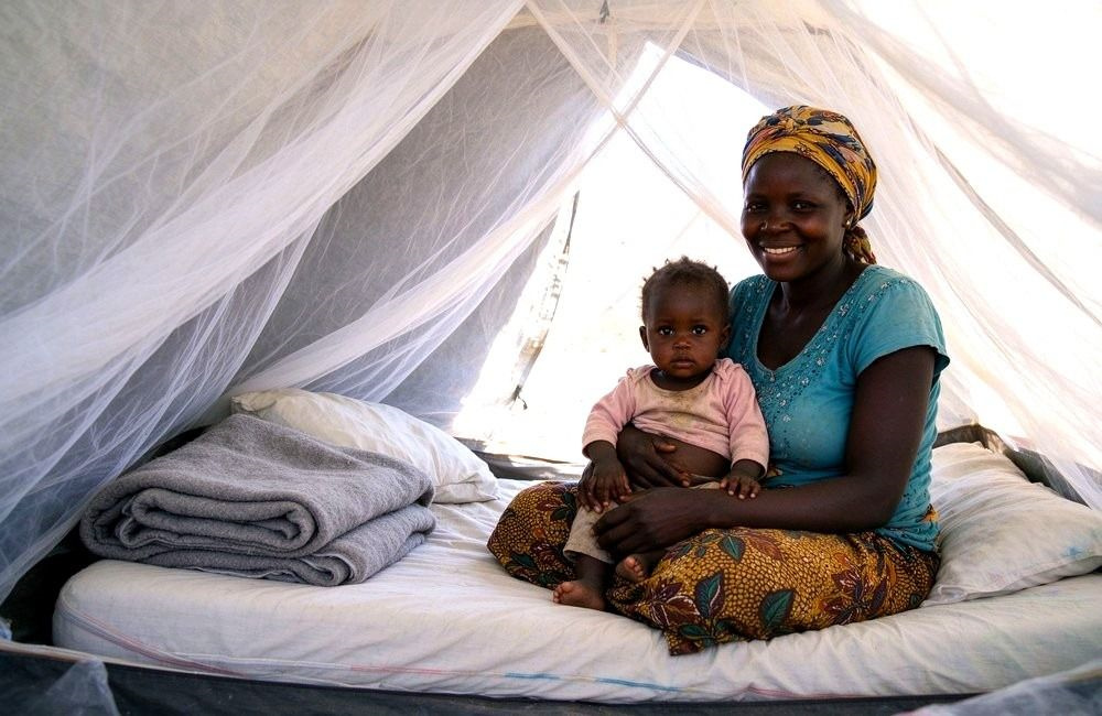 Interior view of an African mother and child inside a flood shelter tent with a clean white mosquito net hanging over the bedding, bright natural light, calm scene